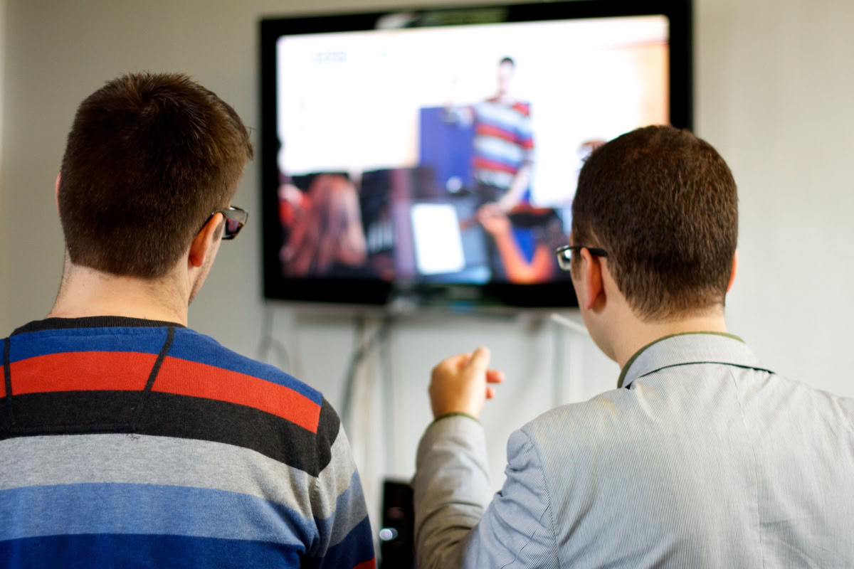 Two people with short hair, seen from behind, watching a television screen.